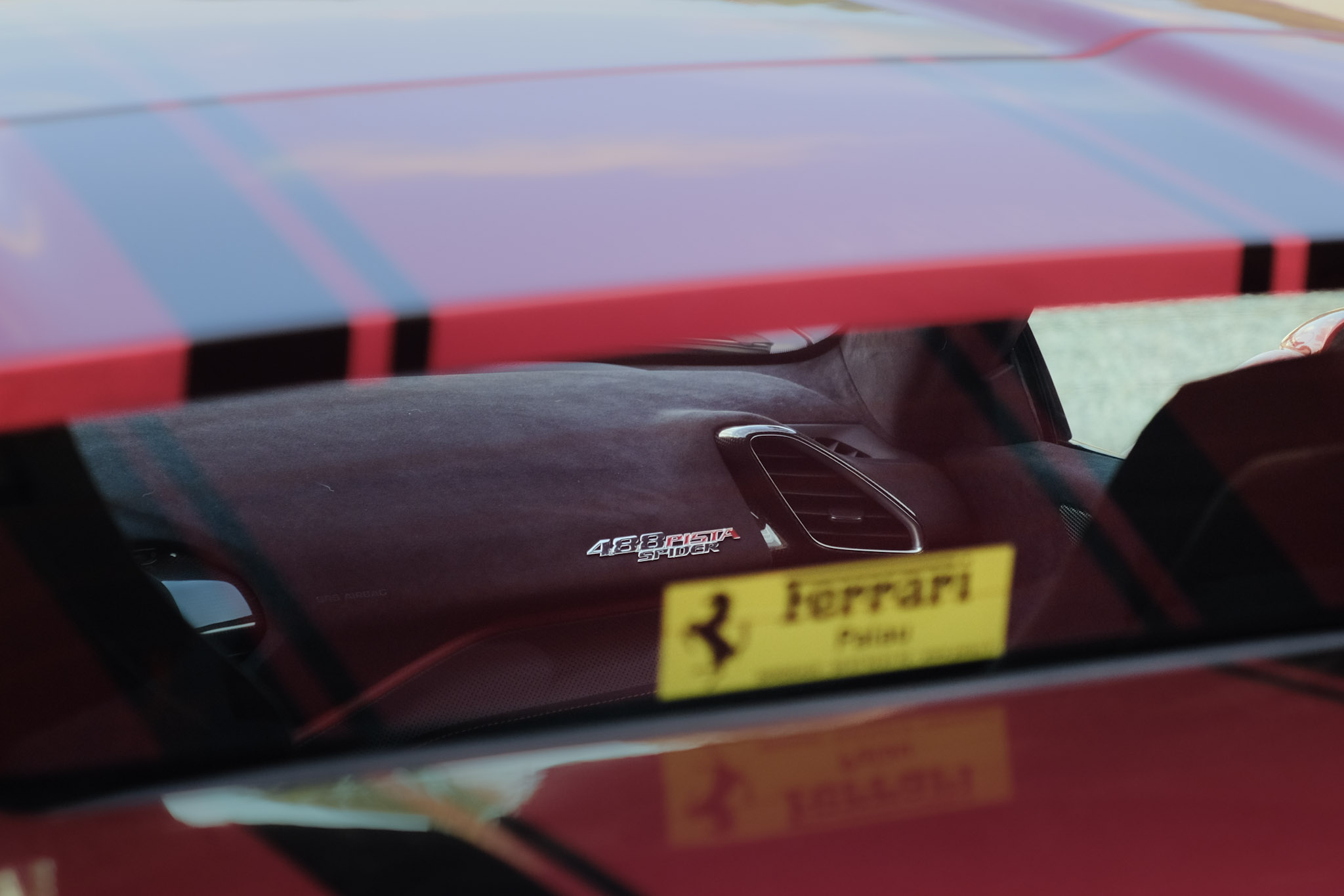 Badge 488 PISTA SPIDER sur planche de bord Alcantara bordeaux — reflet de la carrosserie rouge dans la vitre, sticker Ferrari Palau Bordeaux